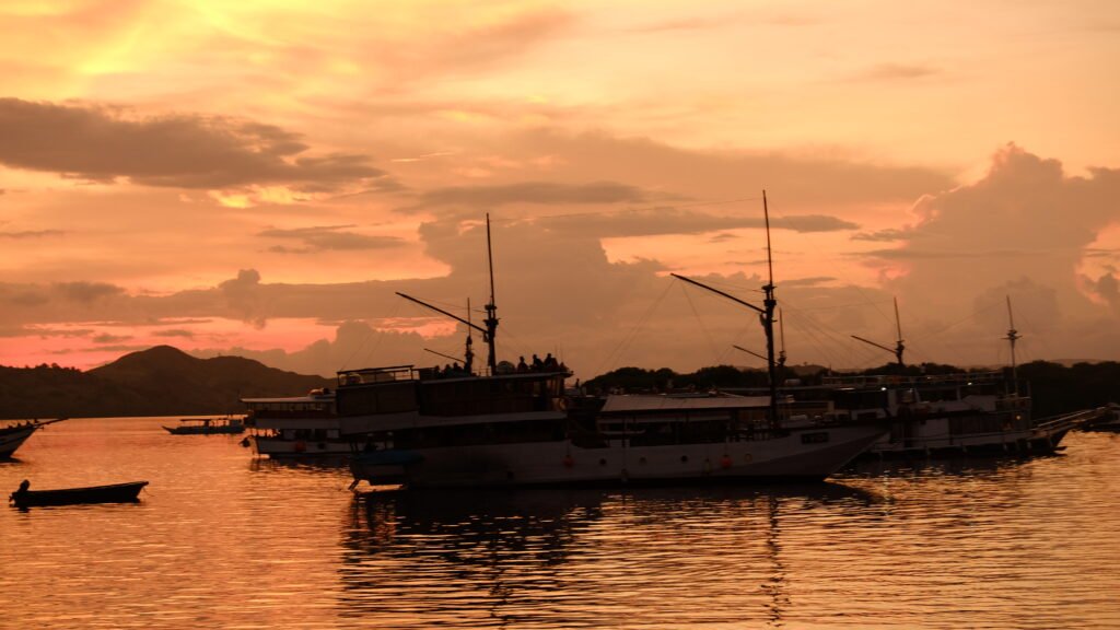 Phinisi boats anchored at Labuan Bajo harbor during a golden sunset with calm sea reflections and dramatic orange sky.