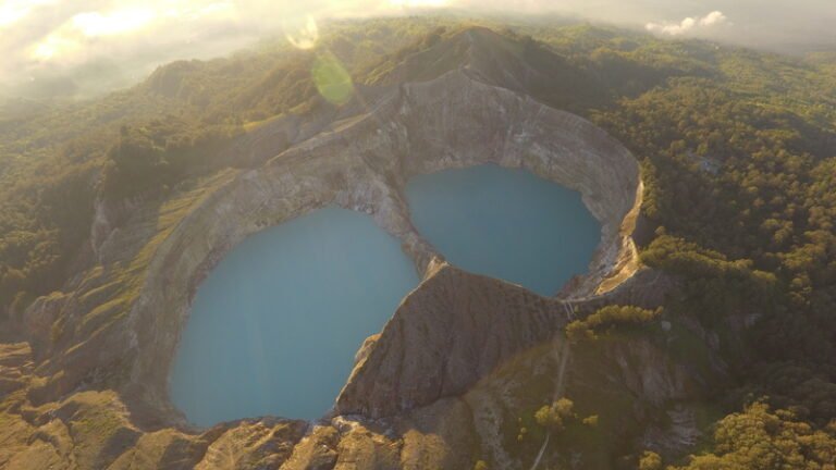 Pemandangan udara Danau Kelimutu dengan dua kawah berwarna biru yang dikelilingi perbukitan hijau saat matahari terbit.