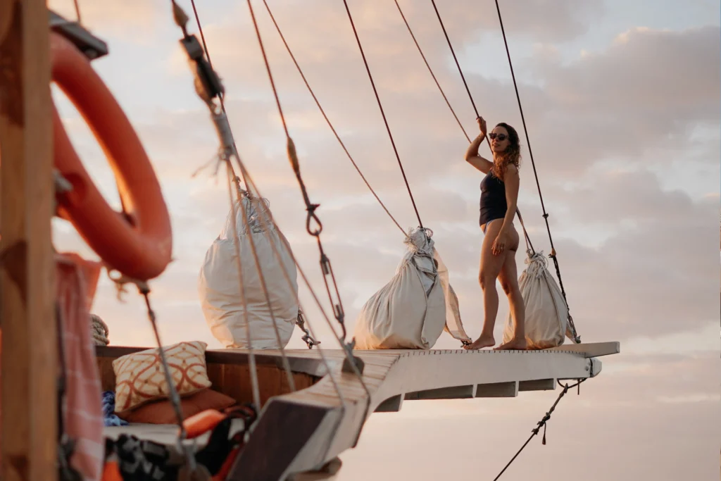 Woman standing on the bow of a luxury phinisi boat at sunset in Komodo National Park, enjoying a scenic sailing experience.