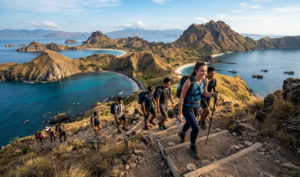 A wide-angle, slightly elevated photograph captures a diverse group of about nine hikers ascending a steep, stepped trail on a sun-drenched, golden-grass-covered hillside on Padar Island, Indonesia. The leading woman in blue top and leggings smiles as she climbs, using hiking poles and carrying a blue backpack and water bottle; behind her follows a line of other trekkers. To the right, a weathered wooden sign with dark, stamped text reads "TRAIL TO PADAR VIEWPOINT." The trail is built from embedded wooden logs and packed earth, winding up the craggy, textured foreground with dry grass and dark rocks. In the background, the stunning, multi-bay panorama of Padar Island unfolds under a bright blue sky with wispy clouds: three distinct turquoise and deep blue bays are separated by rugged, undulating yellow-brown hills. The largest bay features a long crescent white sand beach and a few wooden phinisi sailing boats at anchor. Further in the distance, more rugged islands are visible on the horizon across the sea. The lighting is golden late afternoon sun, casting soft shadows and highlighting the dry landscape and clear water. The composition focuses on the hikers, trail, sign, and the expansive natural landscape.
