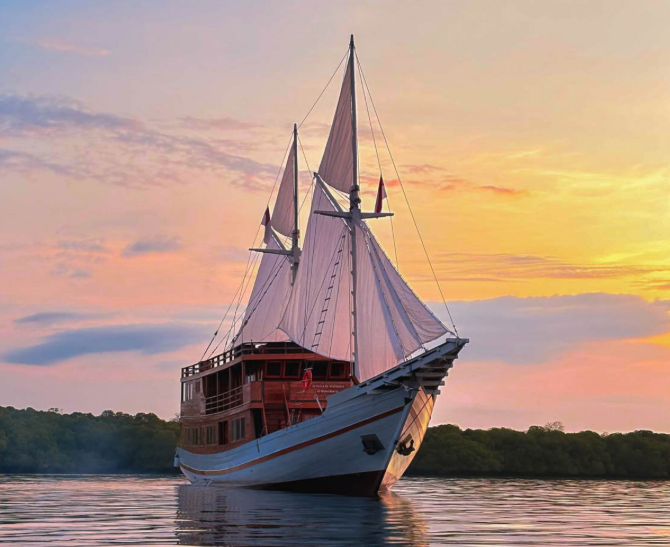 Kapal Phinisi berlayar di Labuan Bajo saat matahari terbenam dengan langit berwarna oranye