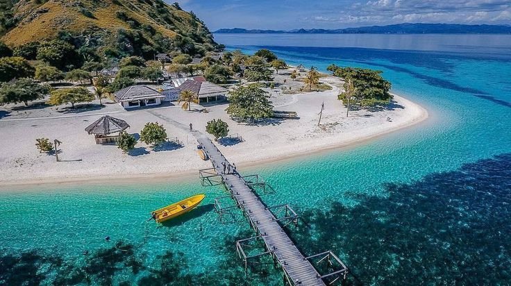 Aerial view of Kanawa Island near Labuan Bajo featuring white sandy beach, wooden jetty, turquoise water, and tropical landscape in Komodo area.”
