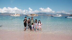 Keluarga berfoto di tepi pantai berair jernih di Labuan Bajo dengan beberapa kapal phinisi berlabuh di laut biru dan latar perbukitan di bawah langit cerah.