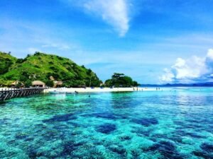 Pemandangan pantai berair jernih di Labuan Bajo dengan pulau hijau dan dermaga, destinasi populer dalam open trip Labuan Bajo untuk snorkeling dan trekking