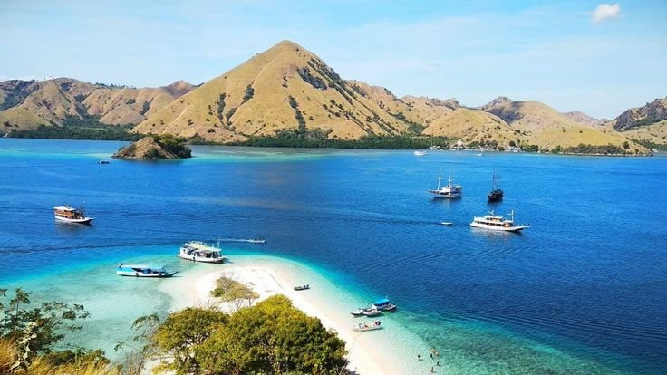 Panoramic view of Labuan Bajo with turquoise waters, white sandbar, boats anchored near a small island, and hills of Komodo National Park in the background.