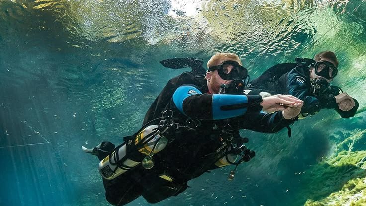 Dua penyelam scuba melakukan latihan buoyancy di bawah air jernih dengan perlengkapan lengkap, menunjukkan teknik kontrol posisi saat menyelam di perairan tropis.