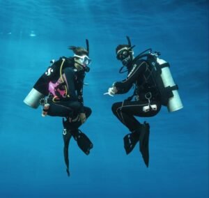 Two scuba divers underwater in full diving gear, communicating with hand signals while hovering in clear blue ocean water.