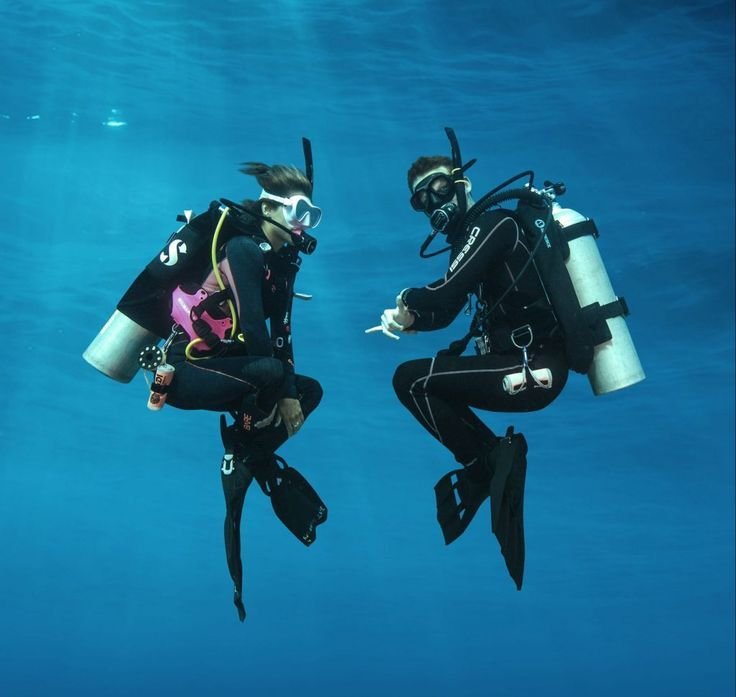 Two scuba divers underwater in full diving gear, communicating with hand signals while hovering in clear blue ocean water.