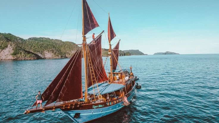 Kapal phinisi berlayar di perairan Raja Ampat dengan layar terkembang dan latar pulau tropis yang indah-visit labuan bajo komodo