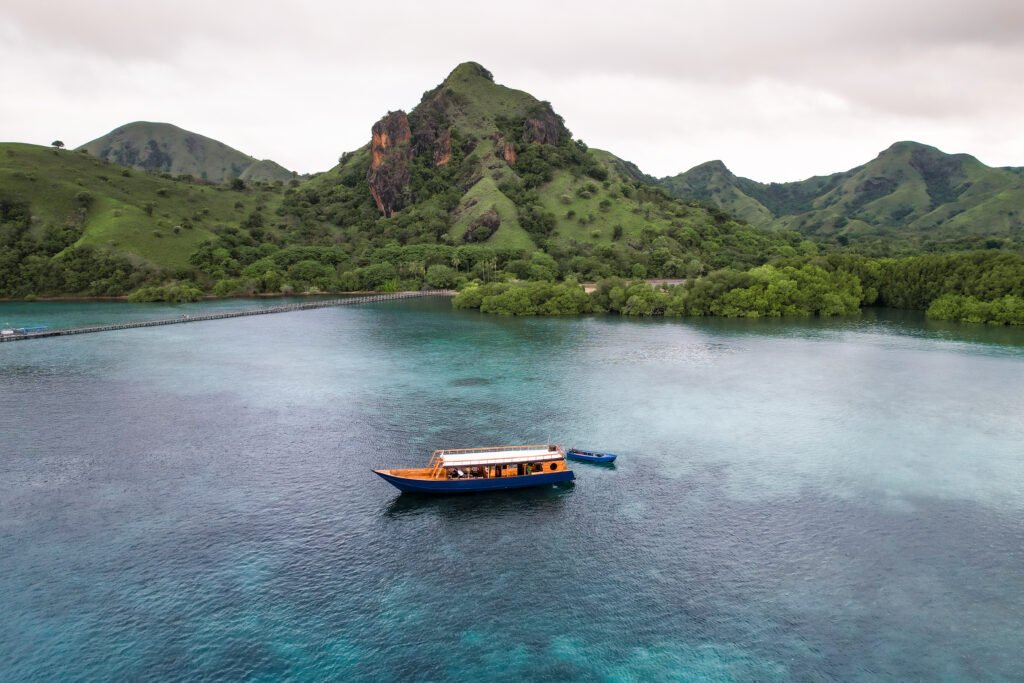 A wooden open-deck boat sailing over clear turquoise waters near lush green hills in Komodo National Park, with a small island and calm tropical scenery in the background.