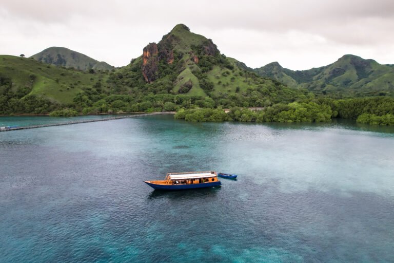A wooden open-deck boat sailing over clear turquoise waters near lush green hills in Komodo National Park, with a small island and calm tropical scenery in the background.