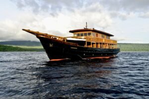Kapal phinisi standar berlayar di perairan Raja Ampat dengan latar laut biru dan pulau hijau tropis-visit labuan bajo komodo