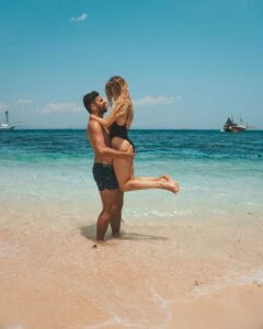 Romantic couple enjoying a tropical beach moment in Labuan Bajo, Komodo National Park, with crystal-clear turquoise water and luxury phinisi boat in the background.