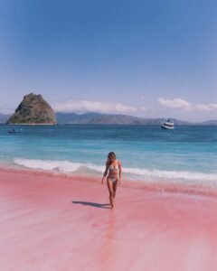 Wisatawan berjalan di pasir merah muda Pink Beach dengan air laut biru jernih dan kapal phinisi di kejauhan bersama Visit Labuan Bajo Komodo
.