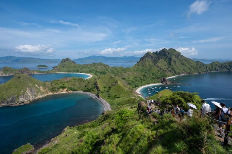 Panoramic view of Padar Island in Komodo National Park with turquoise bays, green hills, hiking trails, and crystal-clear tropical waters under a blue sky.