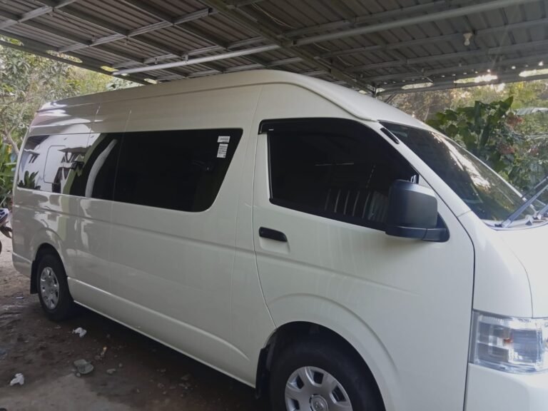 White Toyota Hiace passenger van parked under a covered garage, commonly used for airport transfers, private tours, and group transportation services in Labuan Bajo, Flores, Indonesia.