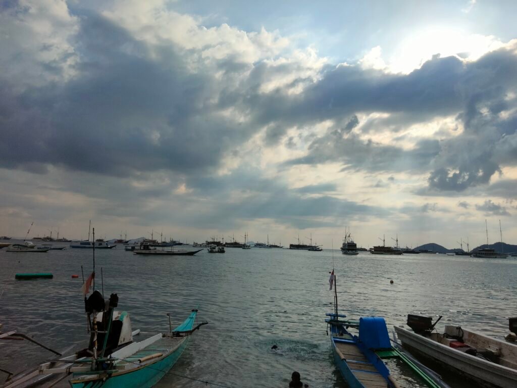 Labuan Bajo Harbor with traditional boats and Phinisi yachts under dramatic cloudy skies, gateway to Komodo National Park, Flores, Indonesia.